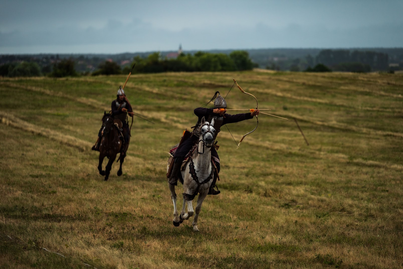 Two archers galloping toward camera, the front rider loosing an arrow, a village spire behind